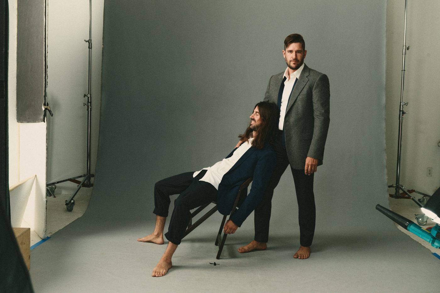 One reclined in a folding chair, the other standing, suits