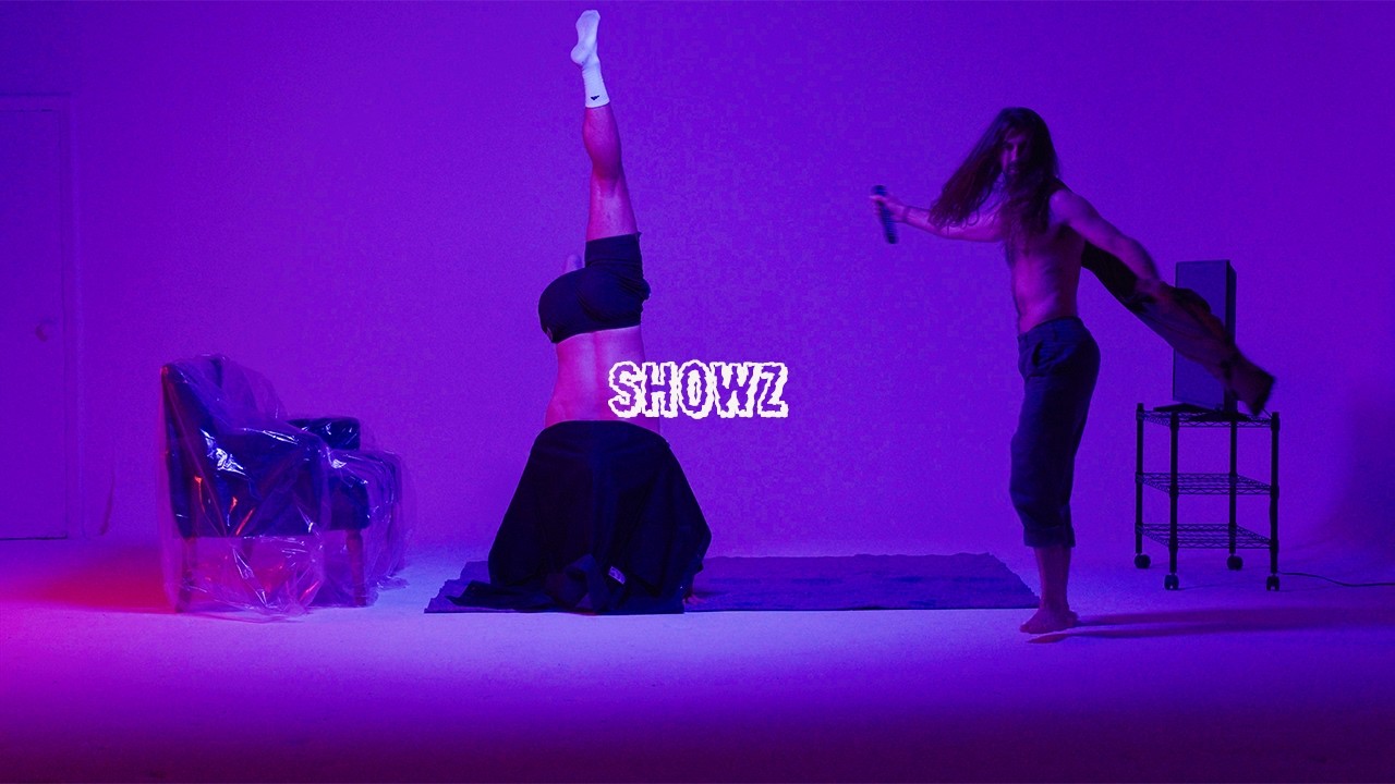 Showz video still: a headstand in tube socks, a man with a microphone, a plastic-wrapped armchair, a TV on a cart, all in purple light