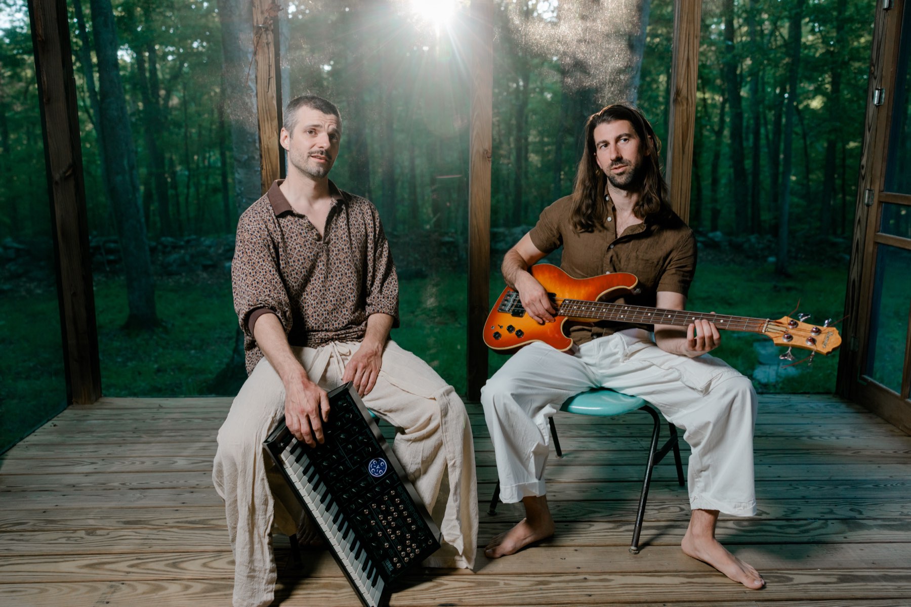 Patrick Manian and Alex Letourneau seated on a screened porch in the woods at golden hour, one with a synthesizer across his lap, one holding a sunburst bass.