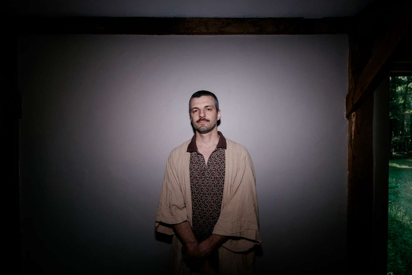 One member alone in a patterned robe under a hard spotlight against a white wall, hands clasped.