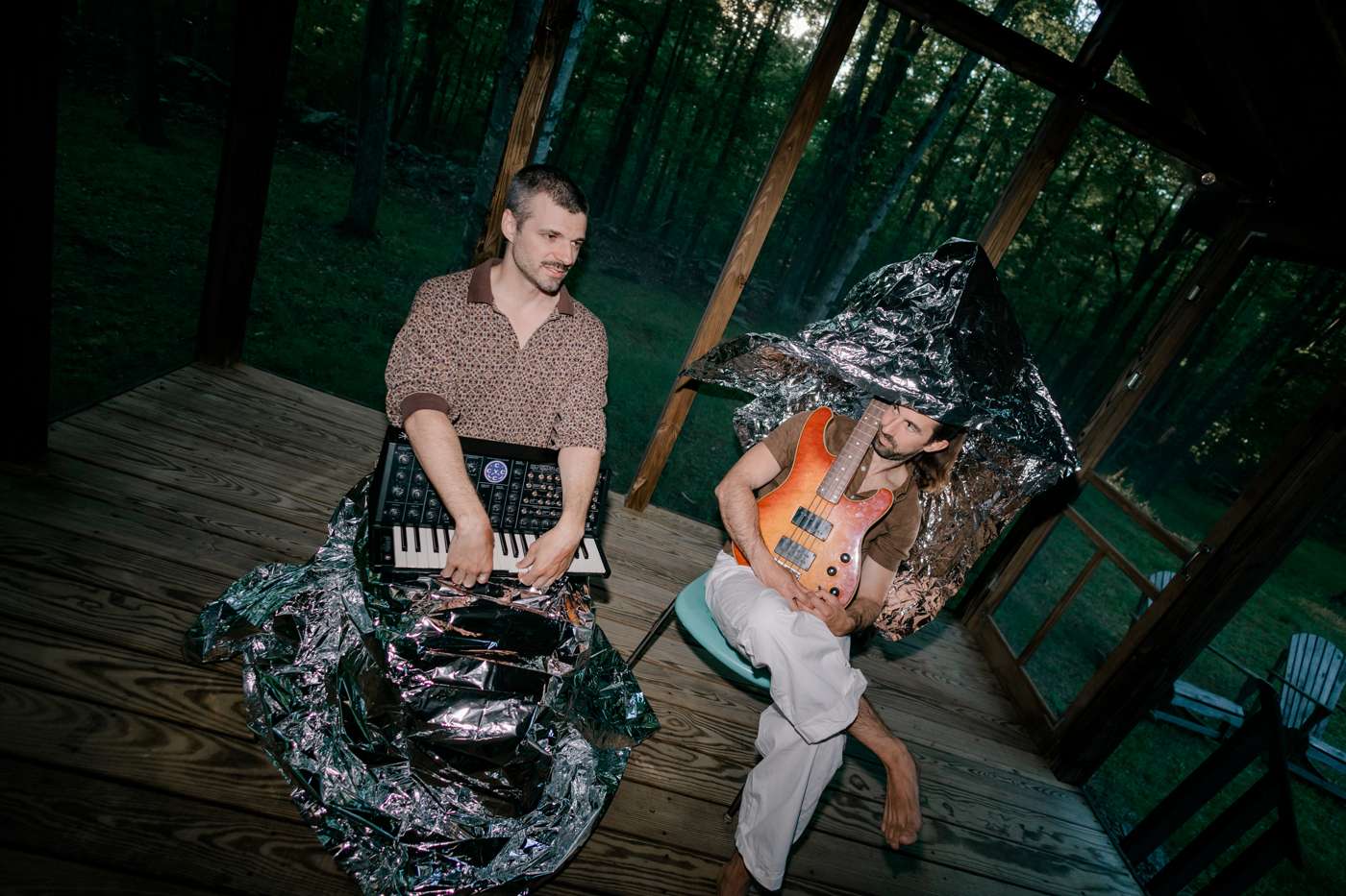 On the porch at dusk, one member flinging a silver emergency blanket into the air, motion-blurred, the other seated behind a synthesizer and a bass.