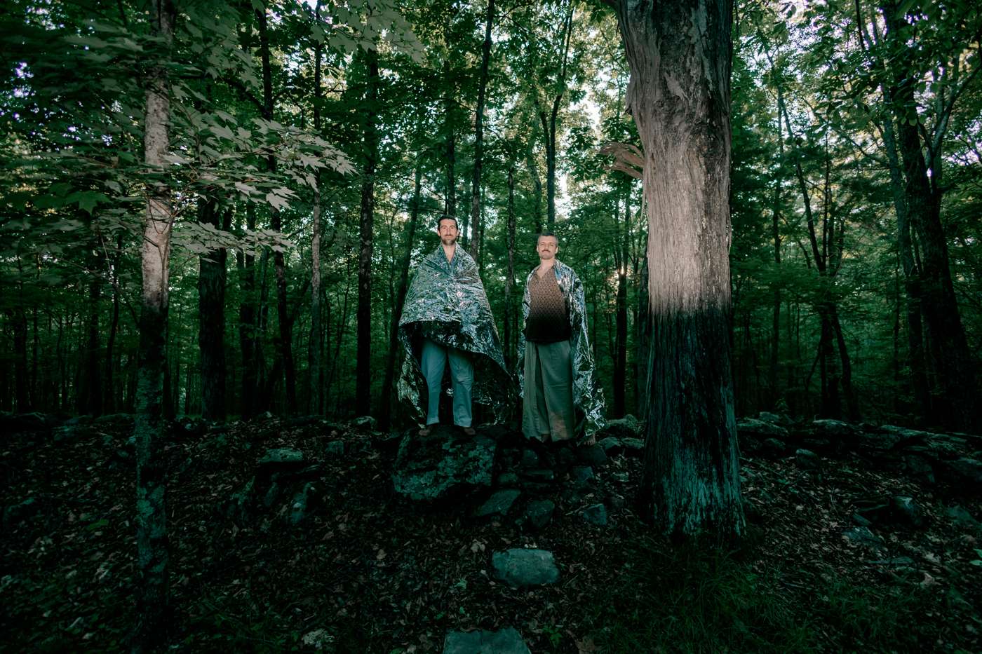 Wide: two small figures under silver blankets on a rock pile deep in green woods.