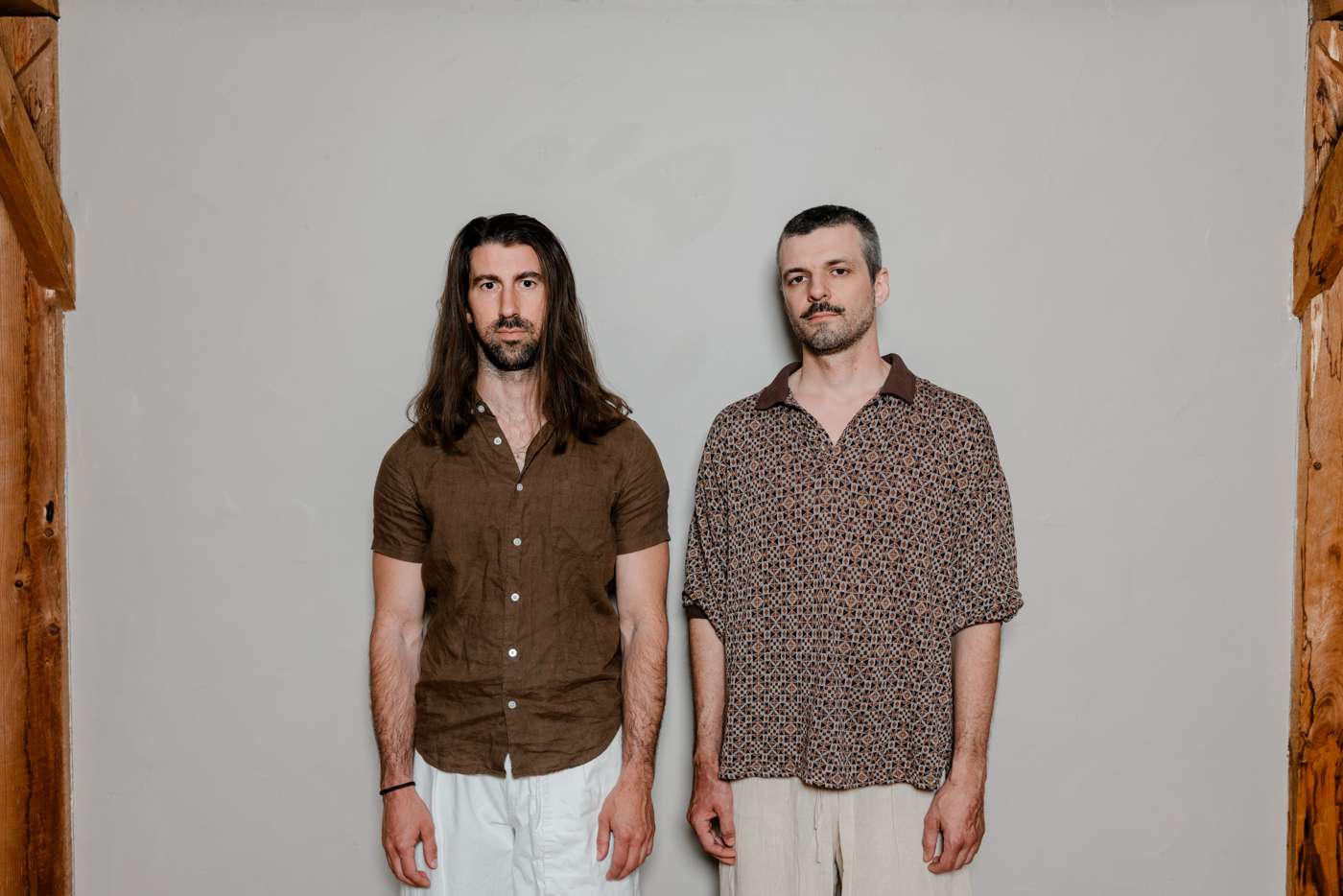 Full-length against a gray wall beside a timber post, deadpan, brown shirts, white trousers.
