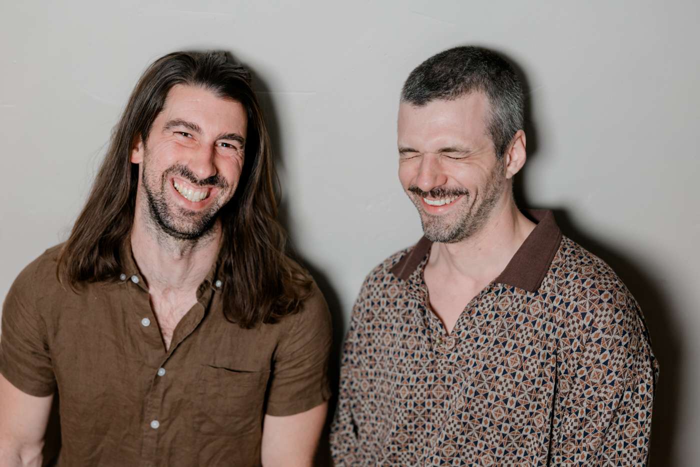 Both members against a gray wall, eyes shut, laughing.