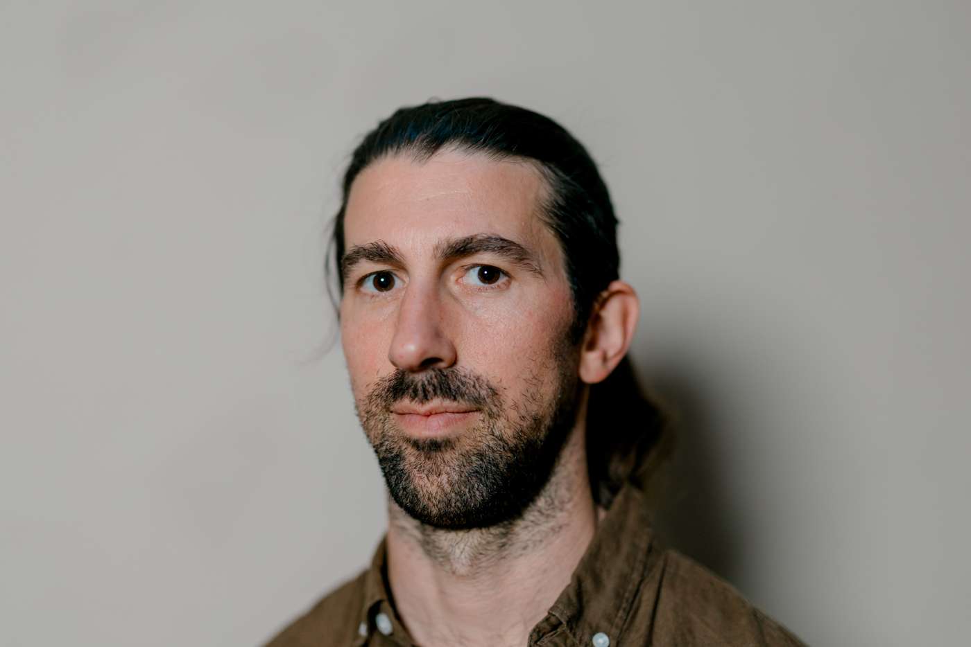 Medium portrait against a gray wall, long hair tied back, brown shirt, looking straight into the lens.