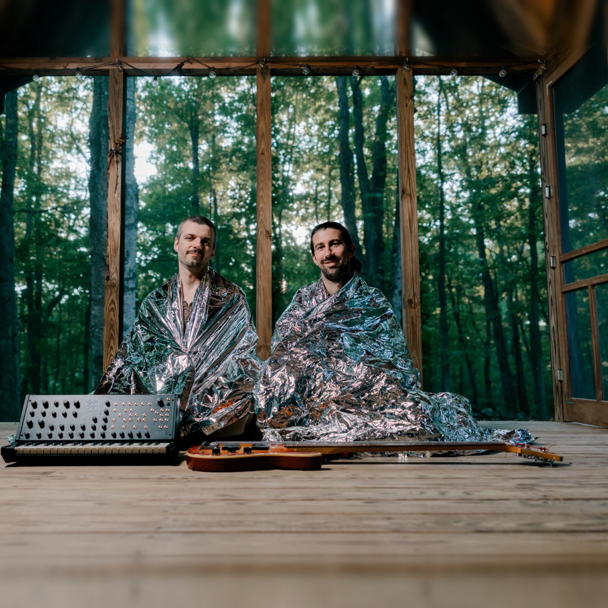 Two men wrapped in silver emergency blankets, sitting on a screened porch in the woods, a synthesizer and a bass laid on the floor in front of them.